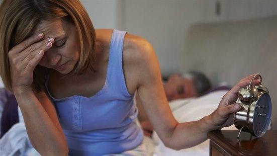 Illustrasjonsfoto: Kvinne sitter på sengekanten. Holder seg for pannen med en hånd, den andre hviler på en vekkerklokke..