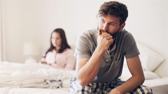 Illustrasjonsfotoet viser et ungt par i sengen. Mannen er i fokus og sitter på sengekanten. Han ser lei seg og frustrert ut. Kvinnen ser man i bakgrunnen, hun sitter i sengen. 
