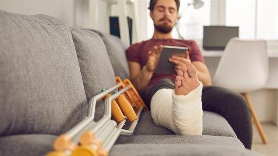 Fotoet viser en ung mann med gips på høyre foten som sitter på en sofa med foten oppå og krykker ved siden av. Han ser på nettbrett.