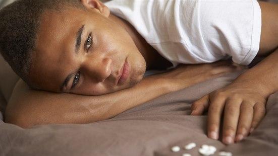 Mørkhudet ung mann ligger med piller foran seg, åpenbart deprimert.