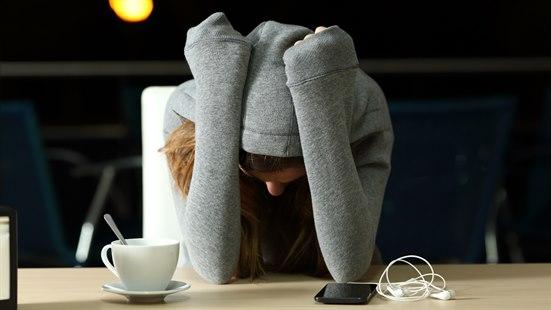 Illustrasjonsfoto: Kvinne med hettegenser støtter albuene mot et bord. Hetten på den grå hettejakken er trukket over hodet, og hun støtter hodet mot armene. På bordet ligger det en kaffekopp og en telefon.