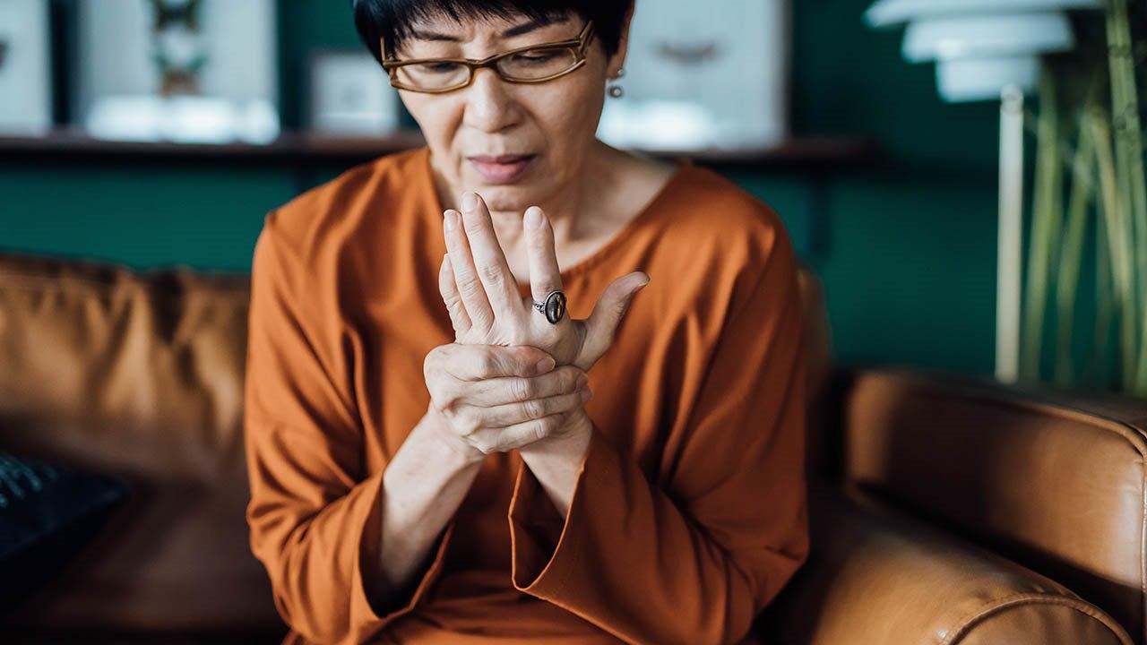 Illustrasjonsfoto av en asiatisk kvinne som sitter i sofaen og holder hånden sin, tydelig viser tegn til håndsmerter.