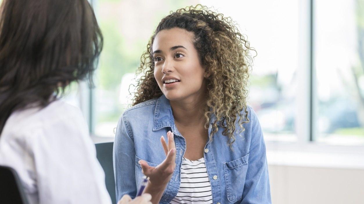Illustrasjonsbilde viser en ung kvinne som er hos legen. Vi ser kvinnen i fokus mens hun forkklarer symptomene sine.