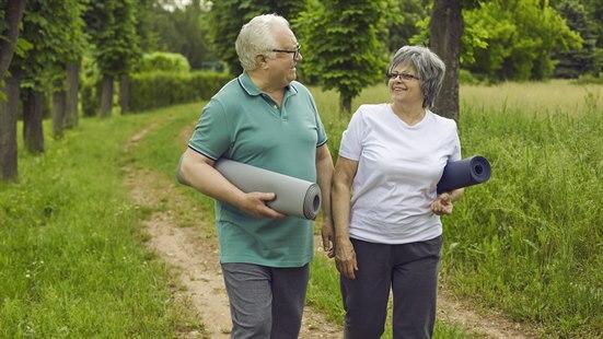 Illustrasjonsfoto av et eldre, smilende par som går på en sti med hver sin yogamatte under arma.
