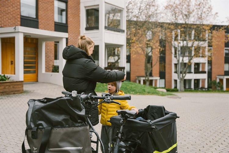Fotoet viser lege Randi etter hun har hentet sønnen sin på barnehagen. Hun står ved siden av sykkelen sin men hun hjelper sønnen å ta på sykkelhjelm