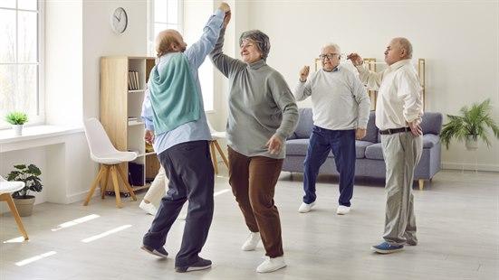 Fotoet viser fire eldre personer (én kvinne og tre menn) som danser i et større rom i en privatbolig eller på et helse- og velferdssenter. 