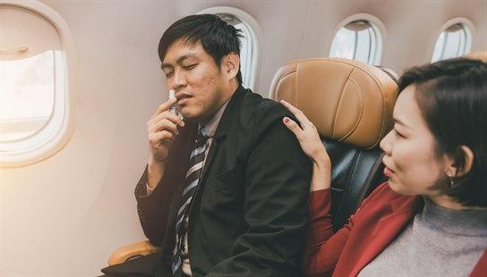 Illustrasjonsfoto av asiatisk mann og kvinne som sitter i hver sitt flysete, der mannen holder en nesespray ved nesen.