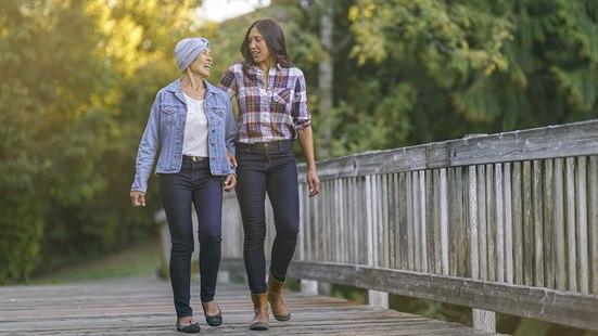 Illustrasjonsfoto av to smilende kvinner som går over en gangbro med grønn skog i bakgrunnen. Ene kvinnen bruker hodeplagg for å skjule hårtap.