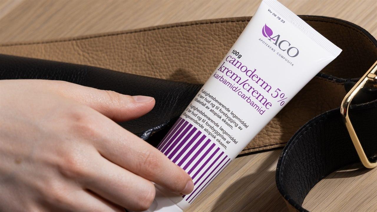 Illustrasjonsbilde viser en kremtube med Canoderm som ligger på en veske. Det ligger en hånd og hviler oppå tuben.