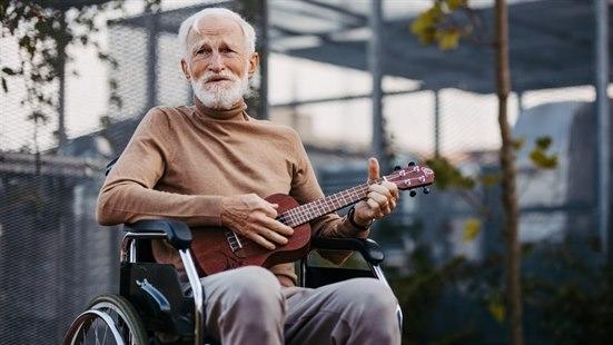 Fotoet viser en eldre slank man ikledd en lysebrun genser. Han sitter på en rullestol ute og spiller på en ukulele. Uteområdet er inngjerdet.