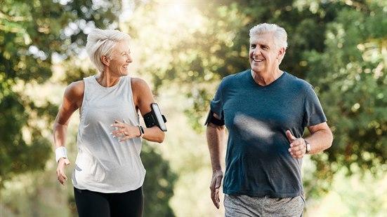 Illustrasjonsfoto av et seniorpar som jogger sammen i skogen. Det er solskinn gjennom trærne og de er smilende og engasjerte mens de nyter en aktiv dag sammen.