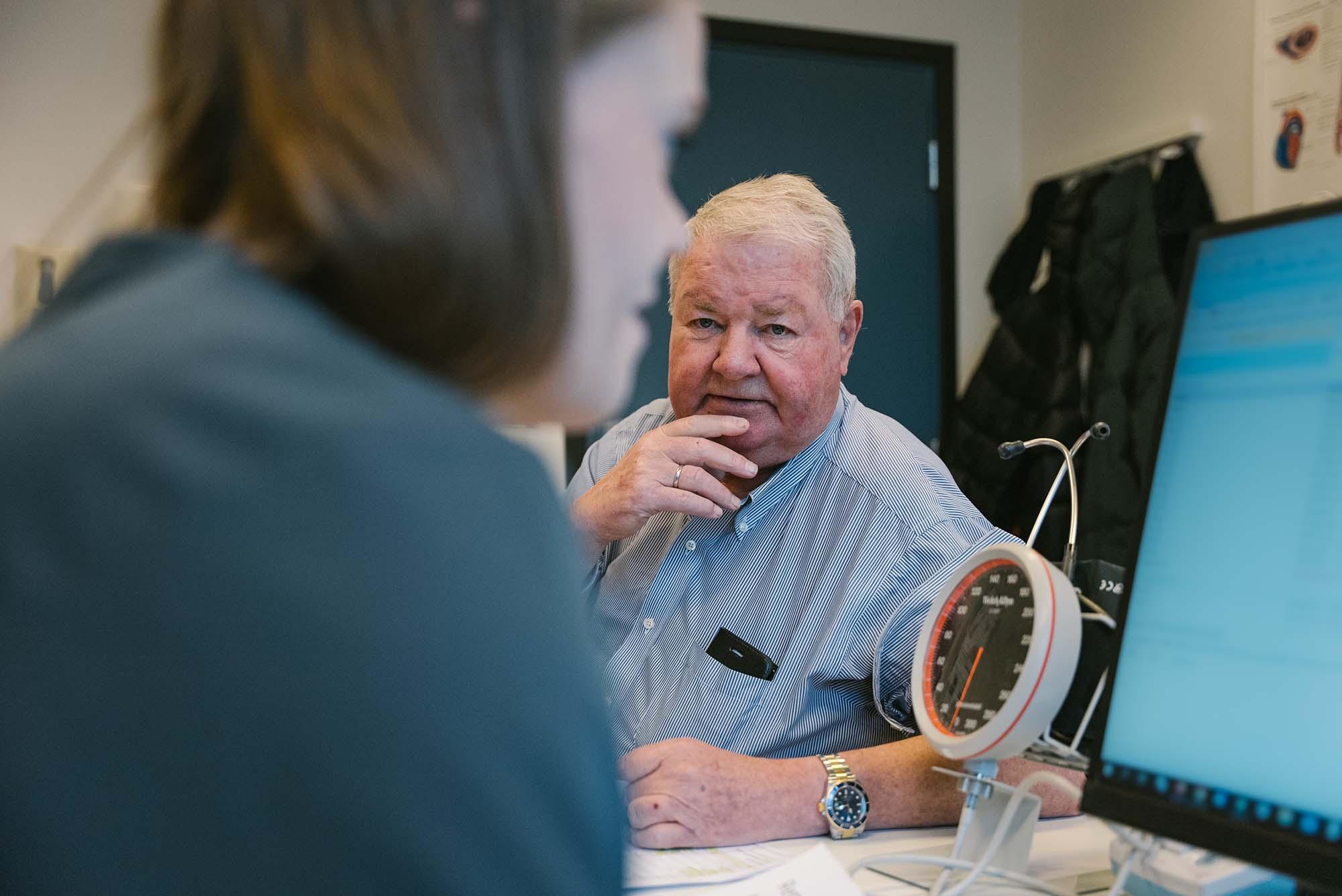 Fotoet viser lege Randi som sitter forran pcen sin på kontoret. I fokus i bildet ser vi pasient John Erik. 