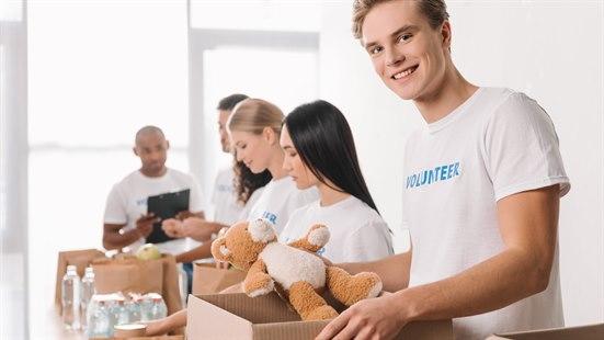 Fem unge mennesker jobber som frivillge og står på rekke og rad og pakker varer i kasser. Den fremeste unge mannen holder en lekebamse i hånda, som skal ned i kassen han pakker. 