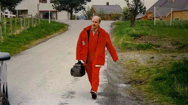 Foto av Terje Johannessen med rød ambulansedrakt og legeveske i hånden, mens han går nedover en vei.