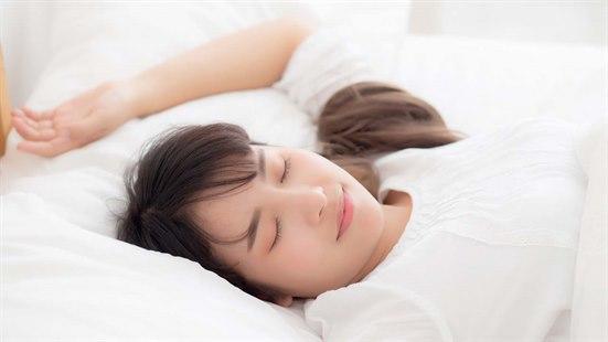En dame som sover på ryggen med en hånd opp. Sengetøyet er hvit.