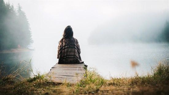 Illustrasjonsfoto av en kvinne som sitter stille på en brygge og mediterer, med blikket rettet mot en rolig, tåkete innsjø.
