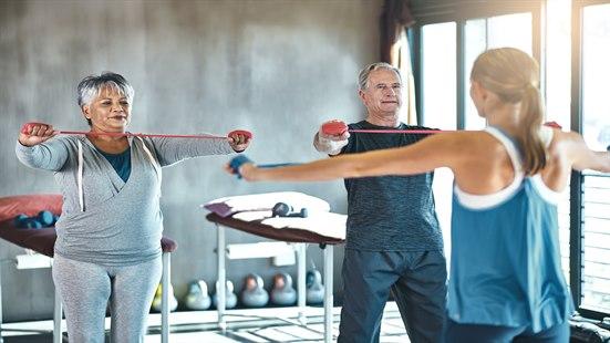 En eldre mann og en eldre kvinne instruert av en trener, utfører en motstandsøvelse i armer og skuldre.