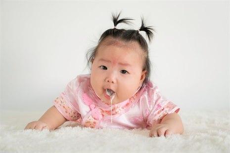 Fotoet viser ansiktet og deler av overkroppen til et 4-måneder gammelt spedbarn av orientalsk opprinnelse ikledd en rosa kjole. Babyen ligger på magen, løfter hodet og gulper opp melk.