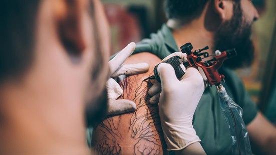 En mann sitter i et tatoveringsstudio , mens tatoveringskunstneren arbeider med å tatovere et tre motiv på armen hans. 