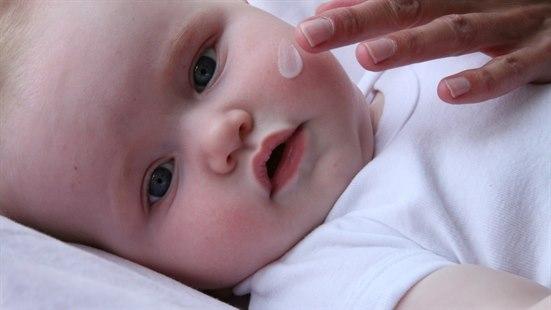 Illustrasjonsfoto av en babys ansikt og en voksens hånd som påfører fuktighetskrem på babyens kinn.
