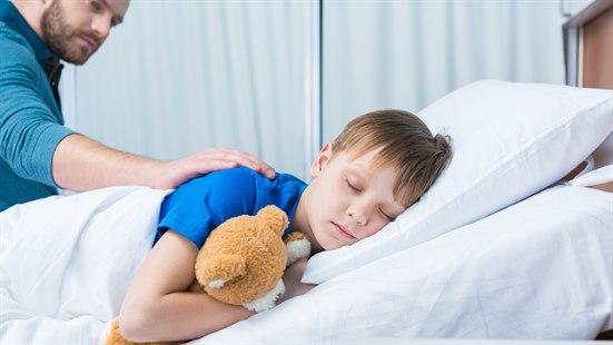 Illustrasjonsfotoet viser en gutt som sover i en sykehusseng. Faren sitter ved siden av og holder en hånd på skulderen hans