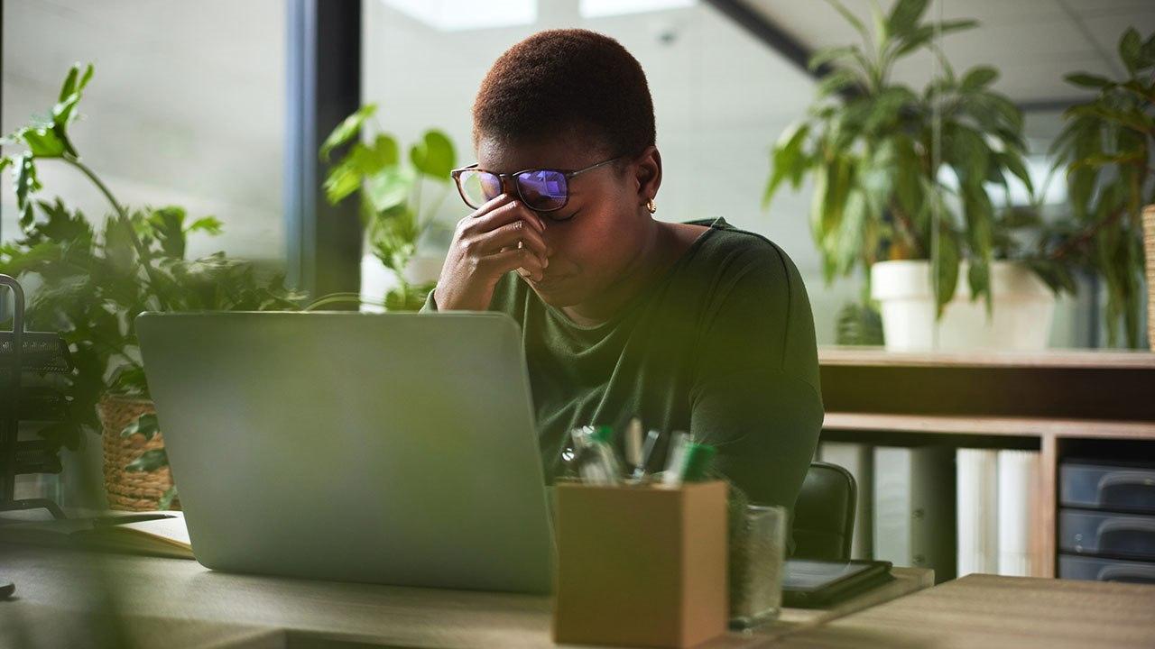 Illustrasjonsfoto av mørkhudet kvinne som sitter i kontorlandskap. Hun holder en hånd over nesen og brillene løftes litt opp. Hun viser tegn til hodepine.