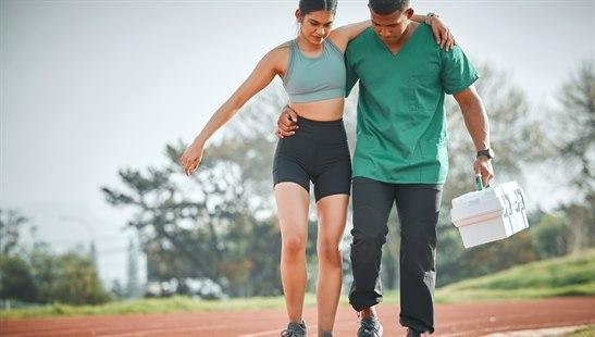 Førstehjelp ved idrettsskade, mann med førstehjelpsskrin som støtter en kvinne ut av et treningsfelt.