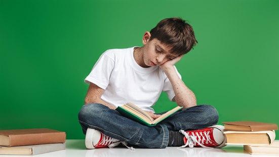 Barn som sitter med bena i kors, en arm støtter hodet og leser i bok.