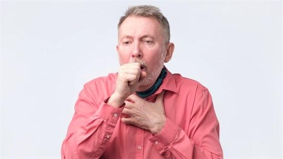 Fotoet viser en godt voksen nord-europeisk mann med grått hår og skjegg som hoster. Han er ikledd en lyserød skjorte. Bakgrunnen er hvit.