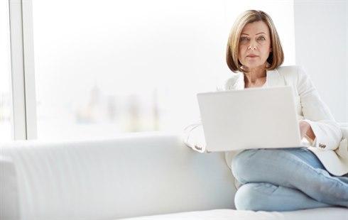 Illustrasjonsfoto viser en middelaldrende kvinne som sitter med bena trukket oppunder seg på en sofa. Hun har en bærbar pc i fanget.