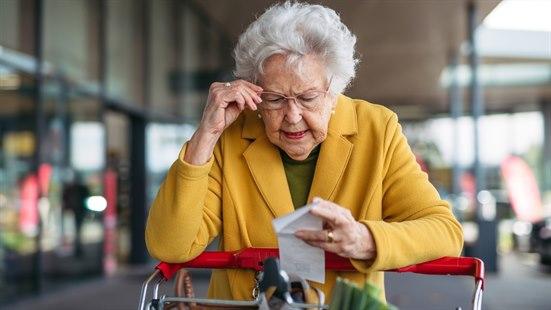 Fotoet viser overkroppen til en eldre dame ikledd en oker kåpe. Hun støtter seg på en handlevogn. Bakgrunnen viser et shoppingsenter. Dama tar på brillene sine og ser på en kvittering, virker litt forbauset.